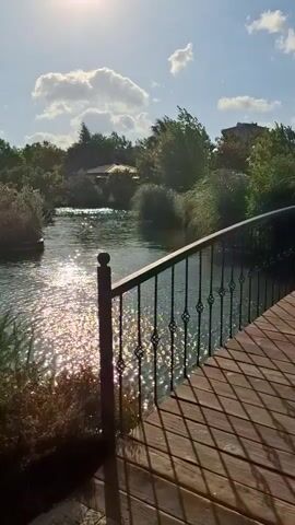 The video shows a scenic view of a body of water, possibly a river or canal, on a sunny day. A wooden bridge with a black metal railing is in the foreground, extending from the right side of the frame. Sunlight glints off the water, creating a shimmering effect. Lush green vegetation, including trees and bushes, lines the banks of the water. The sky is blue with scattered white clouds. In the background, there appear to be buildings or structures partially obscured by the foliage. The perspective suggests the video was filmed from the bridge, looking across the water.