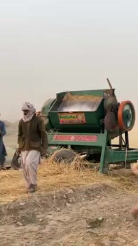 The video depicts an agricultural scene in 'Alaqadari Daman, Afghanistan, focusing on the process of threshing. A group of men, dressed in traditional attire including turbans and shalwar kameez, are actively involved in separating grain from straw.

A prominent green threshing machine with orange and red accents is operating centrally, processing large quantities of dried straw. Text visible on the machine includes "MOHALLAH" and a label stating "MADE IN PAKISTAN." Men are seen manually feeding bundles of straw into the machine. Other workers gather processed straw into piles on the dusty ground, while some are moving material using a wooden tray. Filled and empty sacks, some stacked on a tarp, are also visible.

The scene takes place in a dry, open field with uneven terrain and mounds of earth in the background. The sky is hazy and dust-filled, contributing to a muted, arid atmosphere. The activity appears to be labor-intensive farm work, likely during a harvest or post-harvest period.