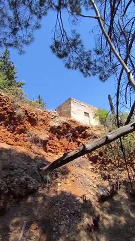 The video shows a steep, rocky hillside covered with dry grass and sparse vegetation. The sky is clear and blue. In the distance, a small, dilapidated building sits on the ridge. The camera pans across the landscape, revealing the rugged terrain and the natural beauty of the area. The overall impression is one of a remote and untamed environment.