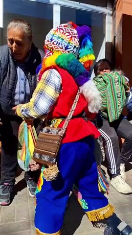 The video shows a person in a colorful, traditional costume, including a brightly colored hat and scarf, standing on a sidewalk. The person is facing a man sitting on a bench. There are posters on a wall behind them. Other people are visible in the background, sitting on benches and walking. The scene appears to be outdoors in a public area, possibly a marketplace or street. The person in costume is wearing a red vest over a plaid shirt, blue pants, and black shoes. The man on the bench is wearing a blue jacket and blue jeans. The overall atmosphere seems festive and possibly cultural.
