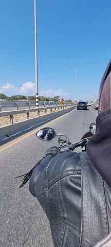 A person wearing a black leather jacket and a motorcycle helmet is riding a motorcycle on a paved highway. The sky is blue with scattered white clouds. There is a metal guardrail to the left of the motorcyclist. Several other vehicles are in front of the motorcycle, including a black Range Rover. The Range Rover has a license plate that is partially obscured.  Streetlights line the median of the highway. In the motorcycle’s mirror, the rider is visible.