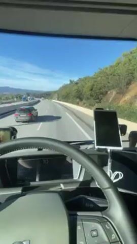 The video shows a first-person view from inside a truck driving on a highway. The truck is in the right lane, and other vehicles are visible on the road, including cars and trucks. The landscape outside the truck window includes green vegetation, hills, and mountains in the distance. The sky is blue with some clouds. Inside the truck cabin, the steering wheel, dashboard, and a phone mounted on the dashboard are visible. The speedometer reads around 70-71. The driver's hands are visible on the steering wheel. The truck seems to be moving at a consistent speed along the highway.