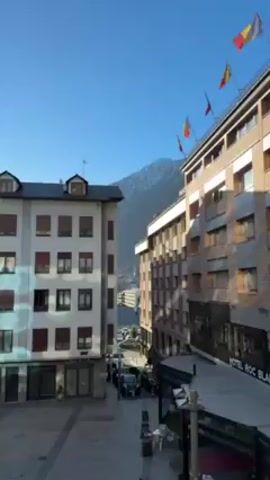 The video shows a view of a town nestled among mountains on a sunny day. The camera is positioned to overlook a small plaza flanked by buildings. The building on the right features flags atop its roof and a sign identifying it as the "Hotel Roc Blanc." Several cars are parked along the street in front of the buildings.  In the distance, a large mountain looms, its peak covered in snow and a tall structure visible at the very top. The sky is a clear, bright blue.