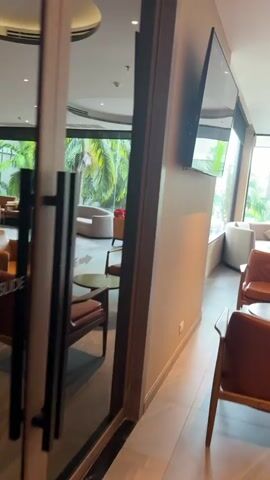 The video pans through a hotel lobby or lounge area. It begins with a focus on chairs and tables, then moves to show large windows overlooking a tropical outdoor area, possibly a pool or garden. The camera continues to pan, revealing more seating arrangements, a large cylindrical column with seating around its base, and a foosball table. In the background, a bar area with lighting is visible, as well as artwork on the walls. The overall impression is of a modern, comfortable, and somewhat upscale public space. There are no people visible in the video.