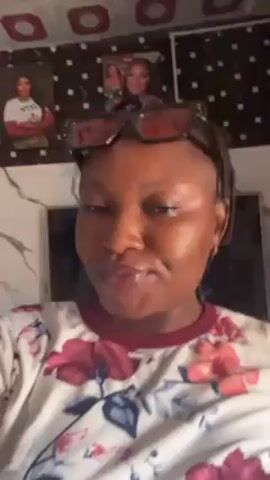 A woman with braids and sunglasses atop her head is recording a selfie-style video. She is wearing a white shirt with large pink floral patterns. The room has a patterned ceiling and framed pictures are visible on the wall behind her. She smiles briefly and moves her head slightly while filming.