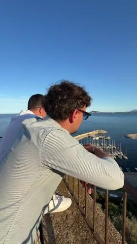 The video begins with a close-up shot of a man leaning on a railing, his back to the camera, looking out at a scenic view. He has dark, curly hair and is wearing a light-colored, striped button-down shirt and sunglasses. His right arm is resting on the railing. To his left, another person, partially visible, is also looking out at the same view. The camera then pans to the left, revealing a wider view of a harbor filled with numerous sailboats and yachts. The water is calm and reflects the clear blue sky. A breakwater juts out into the sea, protecting the harbor. Beyond the harbor, there is a town with buildings nestled among green hills. The sun appears to be setting or rising, casting a warm glow over the landscape. The foreground of the wider shot shows more greenery, suggesting the vantage point is elevated. The overall scene is peaceful and picturesque.