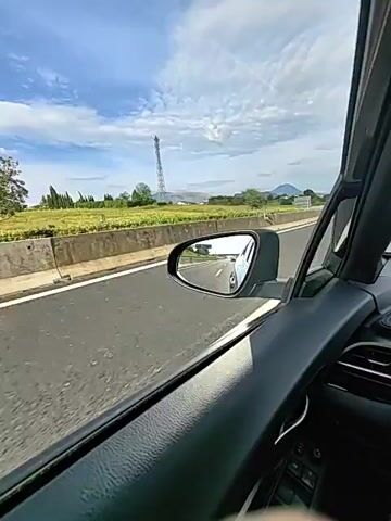 The media file captures a first-person perspective from inside a moving car, looking out the passenger side window. The scene is a highway in Cassino, Italy, with a clear blue sky dotted with white clouds. The time of day appears to be late morning or early afternoon, judging by the bright sunlight casting shadows.

The primary focus is the road, a concrete barrier, and a field of green crops stretching to the horizon. In the distance, rolling hills and mountains are visible. A tall cell tower stands out against the sky to the left of the frame. A white sign with a grid pattern is also visible along the roadside.

The side mirror reflects another vehicle, likely a white car, in the lane behind. The interior of the car is visible in the foreground, including the door panel with window controls and a portion of the dashboard. The overall mood is one of travel and movement, with a sense of open road and scenic vistas. There is no visible text in the media file.