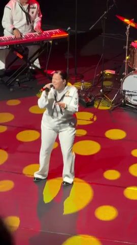 A medium shot shows a woman in a white U.S. Navy uniform singing into a microphone while standing on a stage. The woman has dark hair pulled back and is wearing a white sailor hat with a dark tie. She is wearing white pants and a white jacket with insignia on the sleeves. The stage has a red floor with large yellow circles of light projected on it. In the background, a man in a similar navy uniform is playing a red keyboard. To the right of the keyboard, a drum set is visible. Stage lighting illuminates the scene.