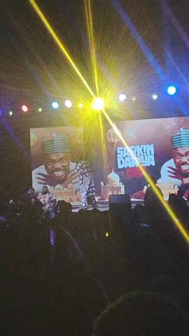 The video shows a live performance in front of a large crowd. A person stands on stage in front of a backdrop displaying the words "SARKIN DARIYA" with images of a man wearing a traditional hat on either side. The stage is lit by several stage lights, including one particularly bright light at the top. There are many people in the audience, and they seem to be facing the stage. The camera is held by someone in the audience, and the view is somewhat obstructed by other people.