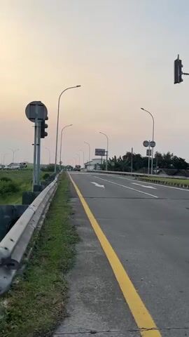 A POV video shows a person riding a bicycle on a highway. The camera pans to the left and right, showing a traffic light, a road with white lines, and green grass. The sky is a soft orange color, indicating either sunrise or sunset. There are cars and buses on the road.