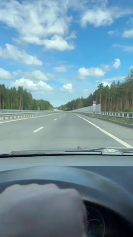 The video is taken from the perspective of a driver inside a car, looking through the windshield. The car is traveling on a highway surrounded by a dense forest of tall pine trees. The sky is blue with scattered white clouds. The highway has multiple lanes with clear white and yellow lane markings. A silver guardrail runs along the side of the road, with a sound barrier visible in some sections. The driver's hand is visible on the steering wheel. The dashboard and a speedometer are partially visible at the bottom of the frame. The car appears to be moving at a moderate speed.