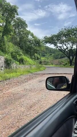 The video shows a view from inside a car driving on a rough, unpaved road with some potholes. To the left, there is a retaining wall with green vegetation growing on top and along the hillside. The road curves to the right, and in the distance, the landscape is covered with lush green trees and hills under a cloudy sky. The side mirror of the car is visible, reflecting a person inside.