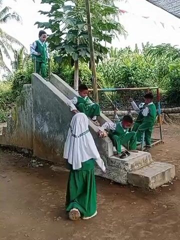 A group of children in green uniforms are playing on a concrete slide. One child is at the top of the slide, two children are sitting on the steps, and another child is at the bottom. A girl in a white hijab and green skirt stands looking at the slide. The slide is located outdoors, with trees and vegetation in the background.