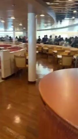 The video shows the interior of a large dining area, likely on a ship or ferry. The space is well-lit with multiple overhead lights. There are many tables and chairs arranged throughout the room. Some tables have red and white checkered bench seating. A central, round, wooden structure is visible in the foreground. In the background, people are seated at tables, appearing to be eating or socializing. The floor is a mix of hardwood and tiled surfaces. Overall, the scene appears to be a bustling and well-maintained dining area.