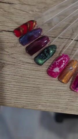 A collection of artificial nails, each affixed to a clear plastic display stick, is arranged in a fan-like pattern on a light-colored wooden surface. The nails showcase a variety of designs and colors, including metallic finishes, marbled patterns, floral motifs, abstract art, and what appears to be text. The lighting is soft and diffused, casting subtle shadows. The background is blurred, suggesting a shallow depth of field.