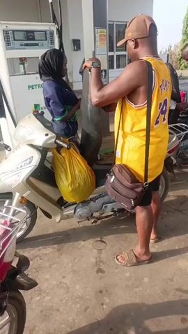 The scene unfolds at a gas station, where a woman wearing a hijab stands beside a gas pump labeled "PETRO." Next to her, a man in a yellow basketball jersey and brown cap interacts with her while holding a yellow plastic bag. The man also carries a brown satchel across his shoulder. The gas station is concrete, with a white and grey structure behind the gas pump. Other individuals and motorcycles are visible in the periphery, suggesting an active gas station environment.