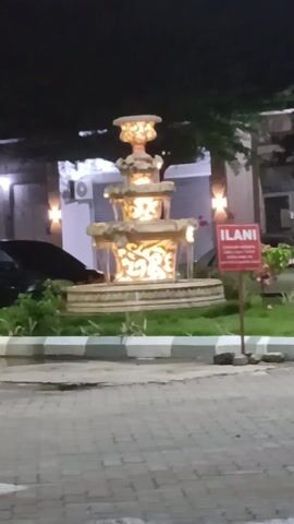 The video shows a three-tiered fountain at night. The fountain is lit from within, highlighting its decorative carvings. A sign reading "ILANI" is next to the fountain. A car is partially visible to the left, and building façades with lights are in the background. The ground is paved.