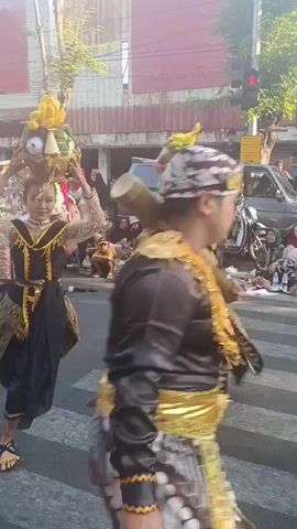 Two young women in traditional ornate black and gold costumes, adorned with sequins and gold trim, are dancing in the street. Each woman balances a woven basket filled with colorful fruits, including bananas and green fruits, on her head. They wear bright green floral hair ornaments. Their costumes consist of a black top with long, shimmering sleeves, a decorative gold sash, and a patterned gold and black skirt. They are performing on a pedestrian crossing in a street with other people watching from the sidewalks and seated on the ground. Behind them, there are parked vehicles and stalls with various goods. The atmosphere appears to be a festive cultural event. The lighting suggests it is daytime.
