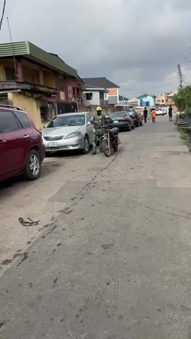 In a medium shot, a man in a camouflage outfit and yellow hat maneuvers a motorcycle down a narrow, gray asphalt street in a residential area. Several cars are parked along the sides of the street, including a burgundy SUV on the left and a silver sedan closer to the center. Further down the street, several people are walking away from the camera. Buildings line both sides of the street, with the building on the left appearing slightly older and more weathered than those on the right. The sky is overcast, casting a slightly muted light over the scene. An object, possibly a broken piece of the motorcycle, lays on the ground near the burgundy SUV.