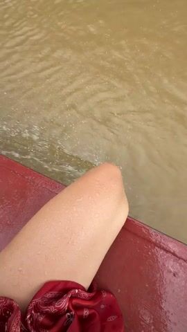A close-up shot shows a person's leg from the knee down, resting on the edge of a red boat. The leg is bare and appears damp, with small water droplets visible on the skin. A red bandana with a paisley pattern is draped over the leg. The water outside the boat is murky brown and appears choppy, with splashes of water hitting the boat's edge near the person's knee. The camera perspective suggests the shot is taken from a slightly elevated, personal viewpoint.