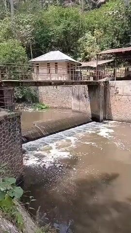 A video shows a small dam on a river. Water flows over the dam, creating white foam. There are stone walls on either side of the dam. A small building with a white roof is located on the dam. A bridge crosses over the dam, connecting to the building. Trees and vegetation are visible on the hillside behind the dam. Two people are standing on a path behind the dam, near some structures. The water appears murky and brown.