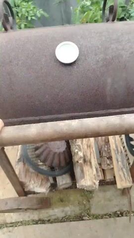 A hand is shown holding onto a horizontal metal bar in the foreground. Behind the bar, a rusty metal barrel with a white thermometer on top is visible. The barrel appears to be part of a barbecue smoker. Inside the barrel, a whole fish wrapped in parchment paper and aluminum foil is placed on a metal rack. The background shows green foliage, possibly tomato plants, and a wooden fence. The overall setting suggests an outdoor cooking environment.