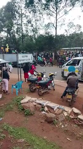 The video shows a scene near Kasimba Primary School, where a crowd of people has gathered. There are numerous motorcycles parked and moving around. A white van with decorations is parked with its door open.  A few small roadside stalls are visible, and the surrounding area includes trees and a grassy patch.  The scene appears bustling with activity.