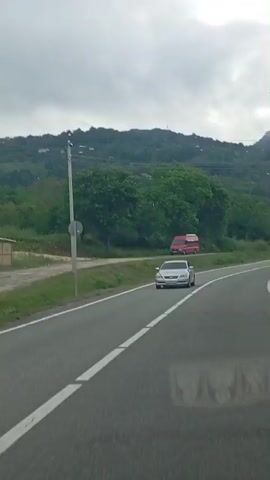 The video shows a view from a vehicle driving on a paved road. The road curves to the right, bordered by a grassy shoulder and trees. In the distance, a hilly, tree-covered landscape is visible under a cloudy sky. Several cars are seen on the road. A silver sedan is in the foreground. Further down the road, a black van and another silver sedan are visible. A red van is parked on the side of the road. There are power lines and streetlights along the road. A small, simple building with a light-colored roof and wooden posts is on the left side of the road near the beginning of the video.