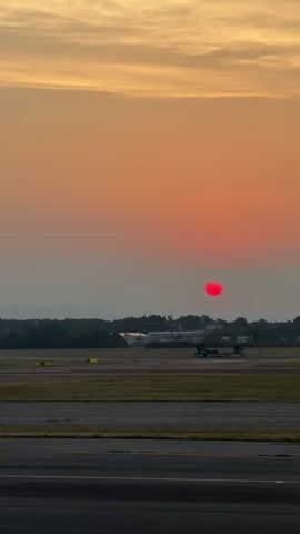 The video shows an airport tarmac at sunset. The sky is a vibrant gradient of orange and yellow, with a bright red sun near the horizon. In the distance, there are trees, buildings, and mountains. On the tarmac, there are several aircraft, including a passenger jet with red and white markings on the left side of the frame. The sun appears to be setting behind the mountains. The video captures a peaceful and serene moment at the airport.