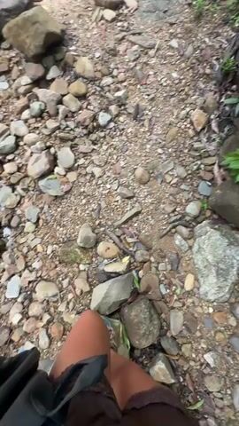 The video shows a person walking on a rocky, unpaved trail. The trail is surrounded by lush green vegetation and large rocks. The person's legs and feet are visible in the foreground, wearing sandals and shorts. The camera perspective suggests the person is walking towards a destination. The trail appears to lead to a waterfall, which is visible in the distance. The waterfall is wide and cascades down a rocky cliff face. There are trees and foliage surrounding the waterfall and the area around it. Several people are visible near the waterfall, some standing and some possibly interacting. There is a small wooden structure, possibly a shelter or rest area, located to the right of the waterfall. The overall environment is natural and appears to be a park or hiking area. The lighting suggests it is daytime, with some dappled sunlight filtering through the trees.