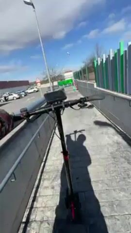 The video shows a person riding an electric scooter along a paved path on a sunny day. The scooter is black with red accents and has a small screen mounted on the handlebars. The person is wearing gloves. The path is bordered by a gray concrete wall on one side and a parking lot on the other. In the background, a Mercadona supermarket is visible. The sky is blue with scattered clouds. The camera is mounted on the scooter, providing a first-person perspective of the ride.