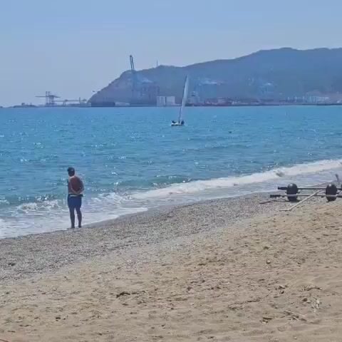 The video shows a beach with a person standing at the edge of the water, facing away from the camera. In the background, a small sailboat with two people is moving across the blue sea. Beyond the sea, a coastal city with industrial buildings and a mountain is visible. The water is choppy, and waves are breaking on the pebbled shore.