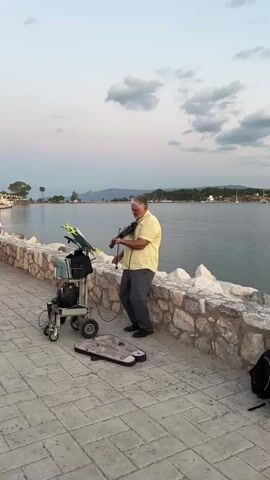 A man is playing the violin on a paved walkway next to a stone wall. He is wearing a yellow short-sleeved shirt and gray pants. He is standing to the right of a wheeled cart that holds music sheets and other equipment. A case for the violin is on the ground in front of the cart. The man is facing the water, which is to his right. In the background, there are trees, buildings, and boats. The sky is a light blue with some clouds. The lighting suggests it is either early morning or late evening.