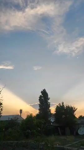The video captures a sunset with crepuscular rays illuminating the sky. The rays emanate from behind clouds and trees, casting beams of light across the sky. In the foreground, there is a stone wall with some greenery growing on it. Behind the wall, there are trees, bushes, and some buildings, including a water tower. The sky is blue with scattered clouds, some of which are illuminated by the setting sun. A small bird is visible in flight.