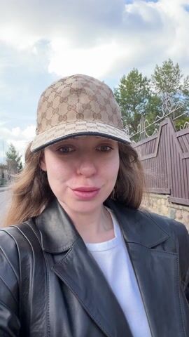 A woman is walking outside on a sunny day. She is wearing a black leather jacket over a white t-shirt, with pink and white striped pants visible below the jacket. She is also wearing a Gucci patterned baseball cap. The woman is looking at the camera and then glances to her right. A wooden fence is visible behind her, and a stone base runs along the bottom of the fence. Green grass and some trees are visible in the background.
