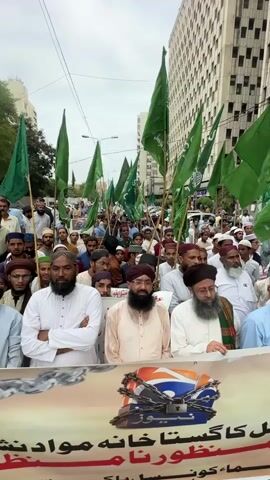A procession of men, predominantly dressed in traditional Islamic attire, march down a street lined with tall buildings, suggesting an urban setting. Many are holding green flags, some with Arabic inscriptions, which are common in Pakistan. The mood appears serious and determined, with many men having stern or focused expressions.

In the foreground, a banner is held by some of the participants. This banner features a logo that resembles a television broadcast symbol, partially obscured by chains and a padlock, suggesting a message about media freedom or censorship. Below the logo, Urdu text is visible.

The scene appears to be taking place during daylight hours, under an overcast sky, indicating potentially mild weather. The architecture of the buildings suggests a modern city, and the presence of the green flags points to a Pakistani context, possibly a protest or demonstration related to religious or political issues. The participants, some with beards and wearing turbans, reinforce the religious undertones of the gathering.