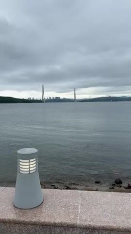 A panning shot moves across a body of water towards a bridge and city skyline under an overcast sky. In the foreground, a modern, grey outdoor lamp with a ribbed white light fixture is positioned on a speckled, polished stone ledge. The water is calm with ripples, and a narrow strip of rocky shoreline is visible between the ledge and the water. In the middle ground, a large cable-stayed bridge with two prominent pylons spans the water. Beyond the bridge, a cityscape with numerous buildings, including several tall, modern structures, is visible on a hilly coastline. The sky is filled with heavy, grey clouds. The camera pans from left to right, revealing more of the bridge and city as it moves.