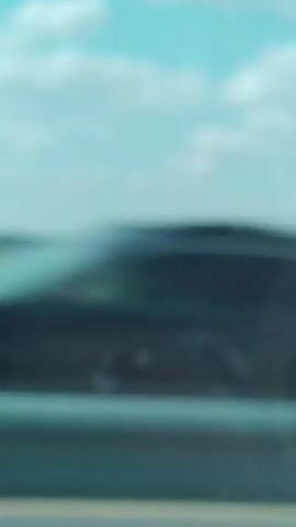 The video shows a blurry view from inside a vehicle looking out a window. The sky is a light blue with some white clouds. Below the sky is a distant, blurry landscape of fields. In the foreground, there is a blurry gray surface, likely the road or highway. At one point, a blurry, dark-colored object, possibly a truck or trailer, passes by on the road. The overall impression is of a fast-moving view of a rural or highway landscape.
