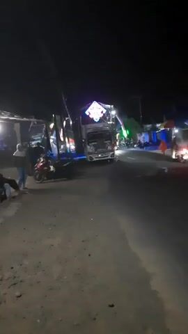 At night, a truck with flashing colorful lights is parked on the side of a road. Several people are standing around the truck and on motorcycles. The road is unpaved and dusty. The scene appears to be a street festival or market.