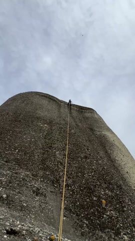 The video shows a person rappelling down the side of a large, gray, rocky outcrop. The person is wearing dark clothing and is attached to a yellow rope. The sky is overcast. The camera pans to reveal a landscape of similar rock formations covered in green trees and vegetation in the valley below. The scene evokes a sense of adventure and the scale of the natural landscape.