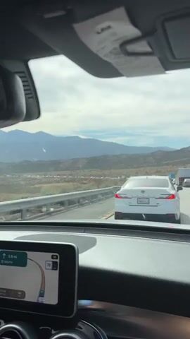 The video shows a view from inside a car, looking out the windshield and side window. The car is on a highway, with a white Toyota Camry in front of it. Mountains are visible in the distance, with a hazy sky. The car appears to be in traffic, with a long line of vehicles stretching ahead. The navigation system on the dashboard shows a route with a speed limit of 70. The terrain outside the car is dry and hilly, with scattered bushes and trees. The sun is shining, casting shadows on the landscape.