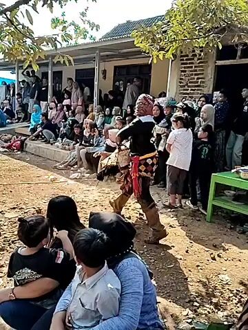 The video opens with a performer in a black outfit and a distinctive mask, possibly part of a traditional dance or performance, moving in a dirt-floored area. Spectators are seated on benches and the ground on the left side of the frame, with a brick building and a covered porch behind them. The performer is holding a large, elaborate prop. The scene appears to be outdoors, with trees and foliage visible in the background. The second shot shows a group of people standing near the entrance of a brick building, some holding up phones to record. The third shot is a close-up of a young man in a black t-shirt, his arms crossed. The fourth shot returns to the performer, who is now moving away from the building and into a more open, wooded area. More people are gathered, and several motorcycles are parked in the background. The final shot shows the performer in the wooded area, with more trees and vegetation. A green table is visible to the right. The overall atmosphere is informal, possibly a village event or celebration.