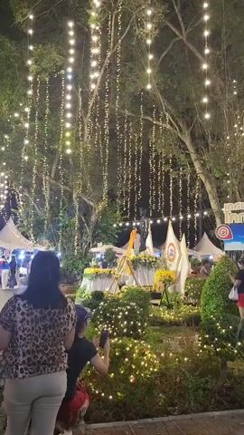 The video pans across an outdoor night scene decorated with numerous strings of white fairy lights hanging from trees. In the foreground, a woman with dark hair stands with her back to the camera, wearing a leopard-print top and white pants. To her right, a child wearing a dark shirt and blue cap is holding a phone, seemingly taking a photo. Behind them, a decorated display with flowers and a golden statue is visible, along with flags. Further back, tents and other people are present. The scene appears to be a festival or market.