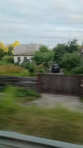 The video shows a blurry, horizontal panning shot of a rural landscape, likely from a moving vehicle. The foreground features a gray metal barrier or guardrail. Beyond that, a wooden fence runs horizontally across the frame. To the left of the fence, there is a dark-roofed structure, possibly a shed. Further back, a white house with a gray roof is visible, surrounded by overgrown grass and bushes. Trees with green foliage are scattered throughout the scene, some illuminated by sunlight. Power lines and a utility pole are also present. The sky above is overcast. The overall impression is one of a somewhat neglected rural area.