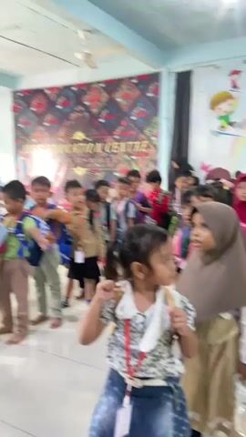 A group of young children, some wearing backpacks and lanyards, are standing in a line indoors. They appear to be in a classroom or event space, indicated by the colorful banner in the background that reads "JESS EDUCATION CENTRE." A woman wearing a hijab stands at the back of the line. The children are looking upwards and to the right, seemingly at something or someone off-camera. The room has a light blue ceiling with a fan and light fixtures. Another banner with cartoon characters and a rainbow is visible on the right wall.