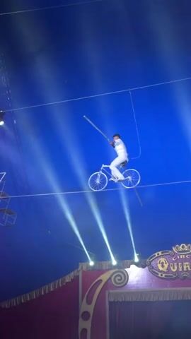A person dressed in a white outfit is riding a bicycle on a tightrope high above a circus tent. The person is holding a long balancing pole. The backdrop is a deep blue, illuminated by bright white and purple spotlights that create beams of light. Below the tightrope, a portion of a circus tent's decorative facade is visible, featuring a sign with golden script. The overall scene depicts a daring circus performance.