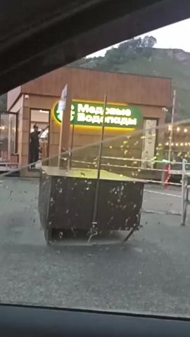 The video shows a scene through a rain-streaked window, presumably from inside a vehicle. In the background, a wooden building with a neon sign reading "Медовые Водопады" (Honey Waterfalls) is visible. A person in dark clothing stands near the entrance of the building. In the foreground, a dark-colored, rectangular object, possibly a dumpster or a storage container on wheels, is positioned in front of the building. There are strings of lights visible to the right of the building. The sky appears overcast, and it is raining, as indicated by the water droplets on the window.