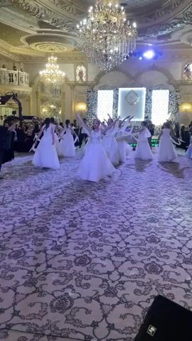 A group of dancers, predominantly female, in white dresses perform synchronized movements in a grand ballroom. A male dancer in a white outfit also participates. The room is ornately decorated with chandeliers, intricate ceiling designs, and a patterned carpet. The dancers move in a line, their arms raised and flowing. The scene appears to be a formal event or celebration.