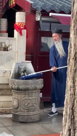 The video depicts an Asian man in traditional clothing standing on a wet pavement, cleaning a stone fountain. He is wearing a long blue robe, white socks, and black shoes, with a black hat and a long white beard. The fountain is adorned with carvings and a lion head spout from which water flows. The man is using a mop-like tool to clean the fountain. Behind him is a red building with a traditional Asian roof. A thick tree trunk is visible to the right, partially obscuring the view. The scene is outdoors in daylight.
