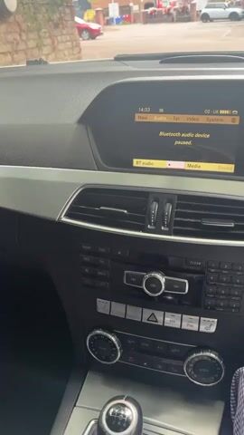 The video shows the interior of a car with a focus on the dashboard. A digital display screen shows the text "Bluetooth audio device paused." Below the screen are car vents and a center console with various buttons and knobs. Part of the steering wheel is visible on the right side of the frame. The view outside the car, seen through the windshield, shows a street with buildings and parked vehicles. The time displayed on the screen is 14:33.